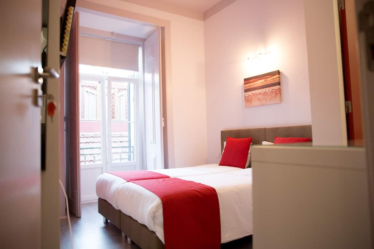 Habitación de hotel con una cama, una colcha roja y blanca, una mesita de noche y una lámpara de pared.