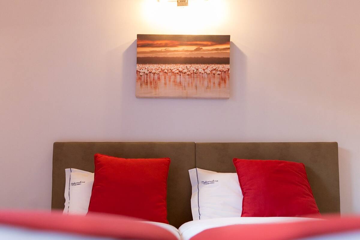 Habitación de hotel con una cama, dos almohadas rojas, una almohada blanca con texto y un cuadro de flamencos en la pared.