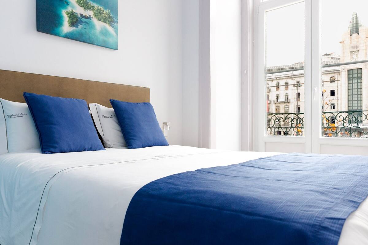 Habitación de hotel con una cama grande, ropa de cama blanca y almohadas azules. Se ve una ventana con vistas a un paisaje urbano.