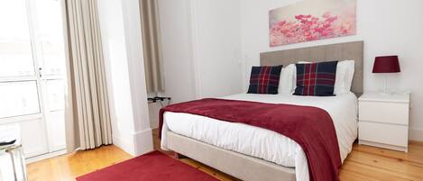 A bedroom with a bed, a nightstand, a lamp, a window with curtains, and a painting on the wall.