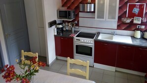 Cocina compacta con microondas, cafetera, estufa y lavaplatos. Hay una silla y una mesa con flores.