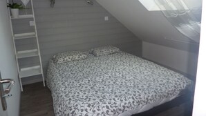 Un dormitorio con techo inclinado, una cama con ropa de cama estampada, un estante blanco con una planta en maceta y una escalera apoyada en la pared.