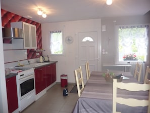 Une cuisine de taille modeste, avec des meubles de rangement rouges, une cuisinière et un évier. On y trouve une table à manger avec des chaises, ainsi qu’une fenêtre agrémentée de rideaux.