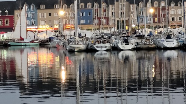 Un puerto deportivo con varias embarcaciones amarradas, reflejadas en aguas tranquilas, y edificios coloridos al fondo.