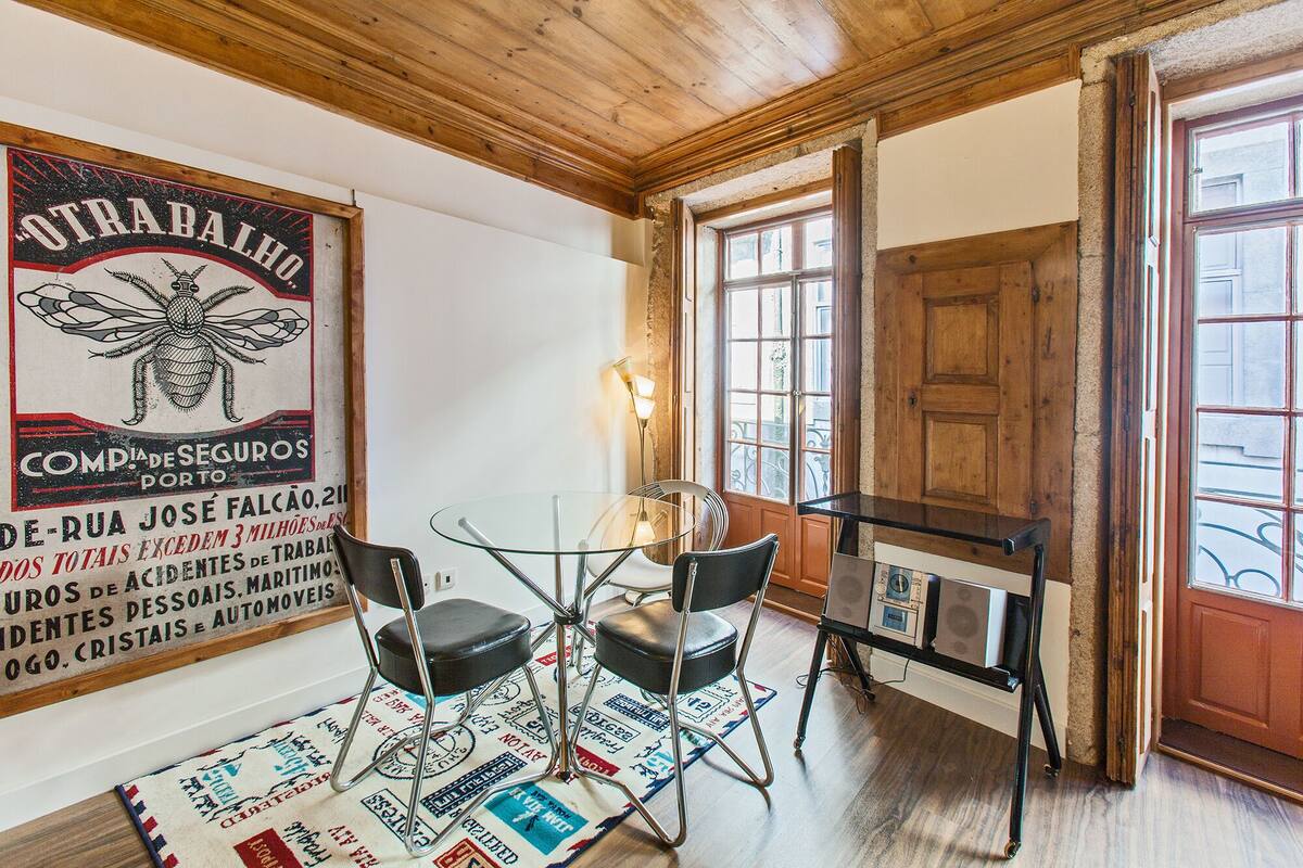 A room with a wooden ceiling, a glass table with chairs, a vintage poster on the wall, and a door leading outside.