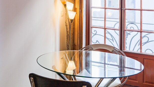 A glass table with chairs, a wall-mounted lamp, and a window with iron railings. - Go2oporto A-Portoments (Porto)