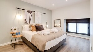 A modern bedroom with a large bed, a bedside table with a lamp, and a window with blinds. - Sweett | Verdi I - Two Bedroom Apartment, Sleeps 5 (Barcelona)