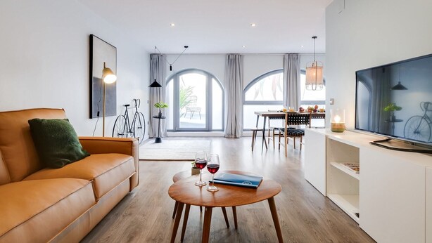 A modern living room with a leather sofa, a wooden coffee table with wine glasses, a dining area with a table and chairs, and a TV on a white stand. - Sweett | Verdi I - Two Bedroom Apartment, Sleeps 5 (Barcelona)