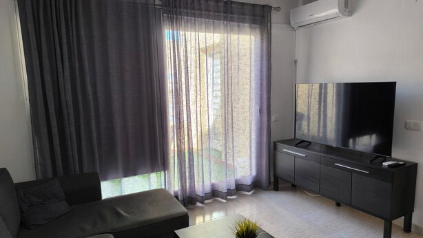 A modern living room with a sofa, a coffee table with a plant, a TV on a cabinet, and a large window with curtains. - Apartment on the beach in Rincón de la Victoria. (Rincón de la Victoria)