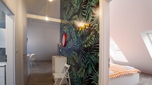Ein modernes Schlafzimmer mit schräg versetzter Decke, einem Bett mit orange-weiß gestreifter Tagesdecke, einem weißen Schreibtisch mit Stuhl, einer kleinen weißen Kommode und einer Wand mit tropischem Blattmuster.