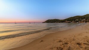 Une plage à l’eau claire, des empreintes de pas dans le sable et, au loin, on aperçoit des voiliers.
