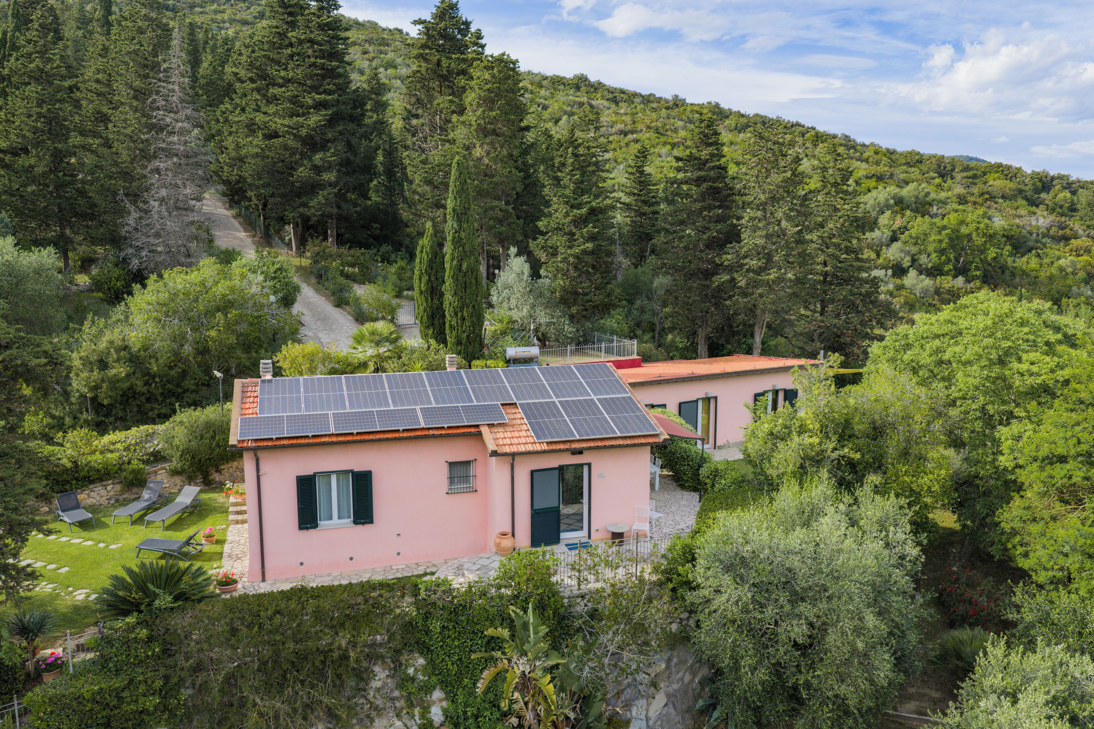 A pink villa with solar panels, surrounded by lush greenery and a mountain backdrop.