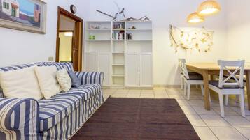 Uma sala de estar com um sofá listrado, uma mesa de jantar com cadeiras, uma estante de livros e um objeto decorativo na parede.