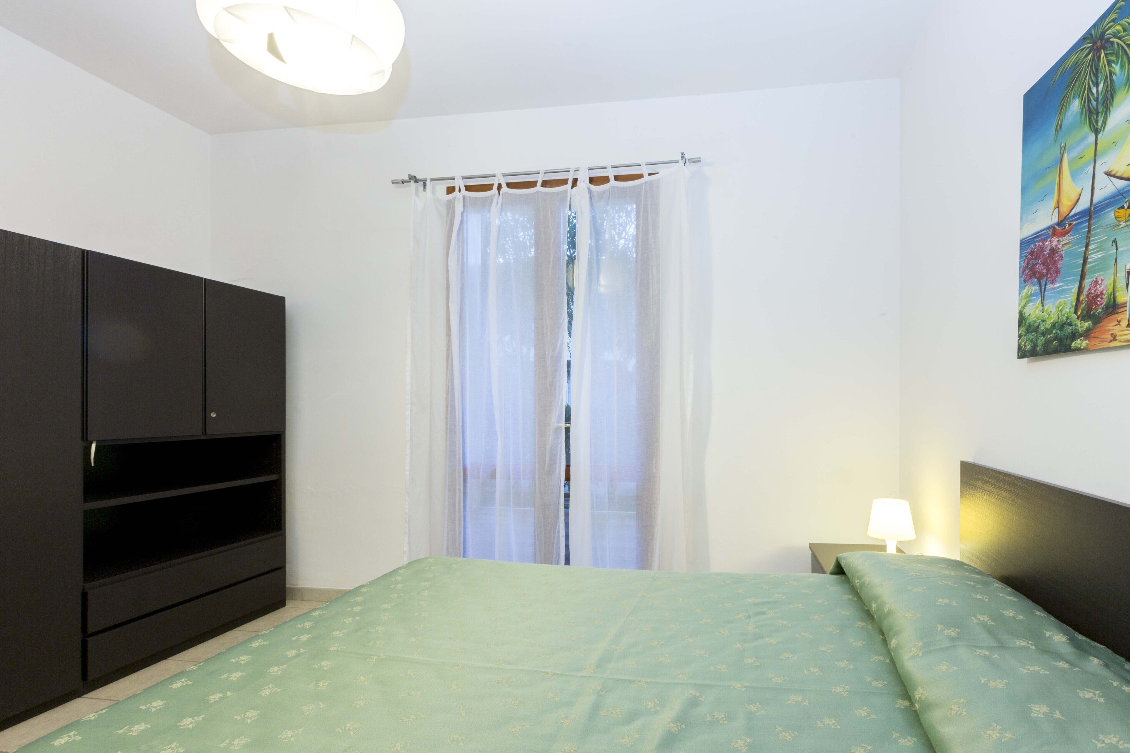 Une chambre à coucher avec un lit, une armoire, une fenêtre avec des rideaux légers et un tableau au mur.