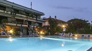 A swimming pool with lounge chairs and umbrellas, surrounded by buildings with balconies and outdoor lighting. - Il Porto Monolocale 4 (Porto Azzurro)