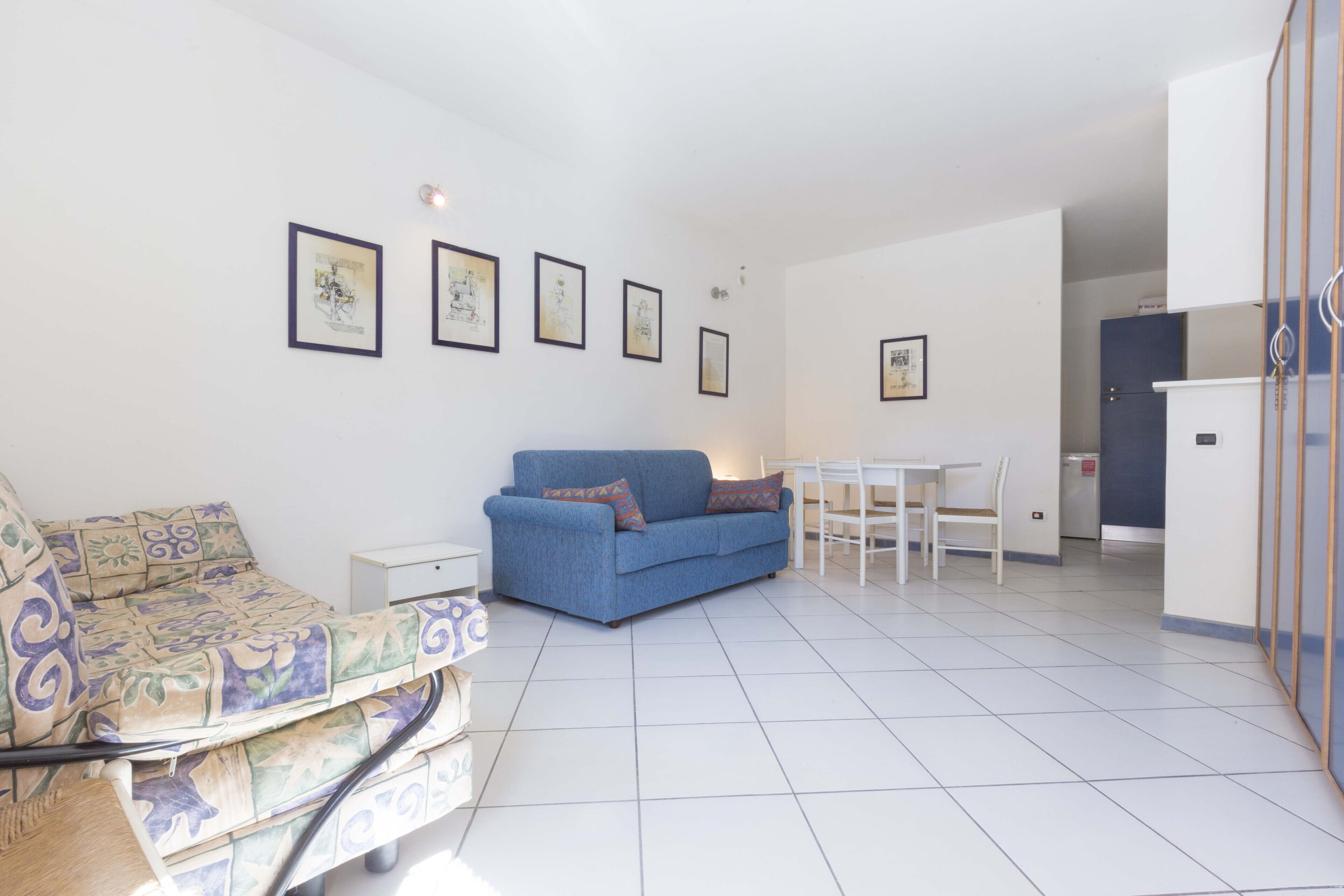 A living room with a blue sofa, a patterned armchair, a dining table with chairs, and framed pictures on the wall.