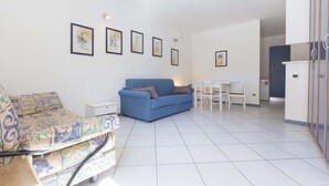 A living room with a blue sofa, a patterned armchair, a dining table with chairs, and framed pictures on the wall. - Il Porto Monolocale 4 (Porto Azzurro)