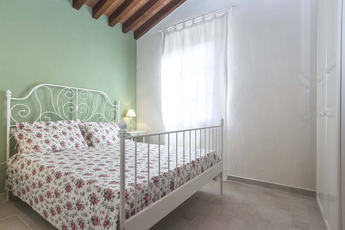A single bed with a floral bedspread, a bedside table with a lamp, and a window with sheer curtains.