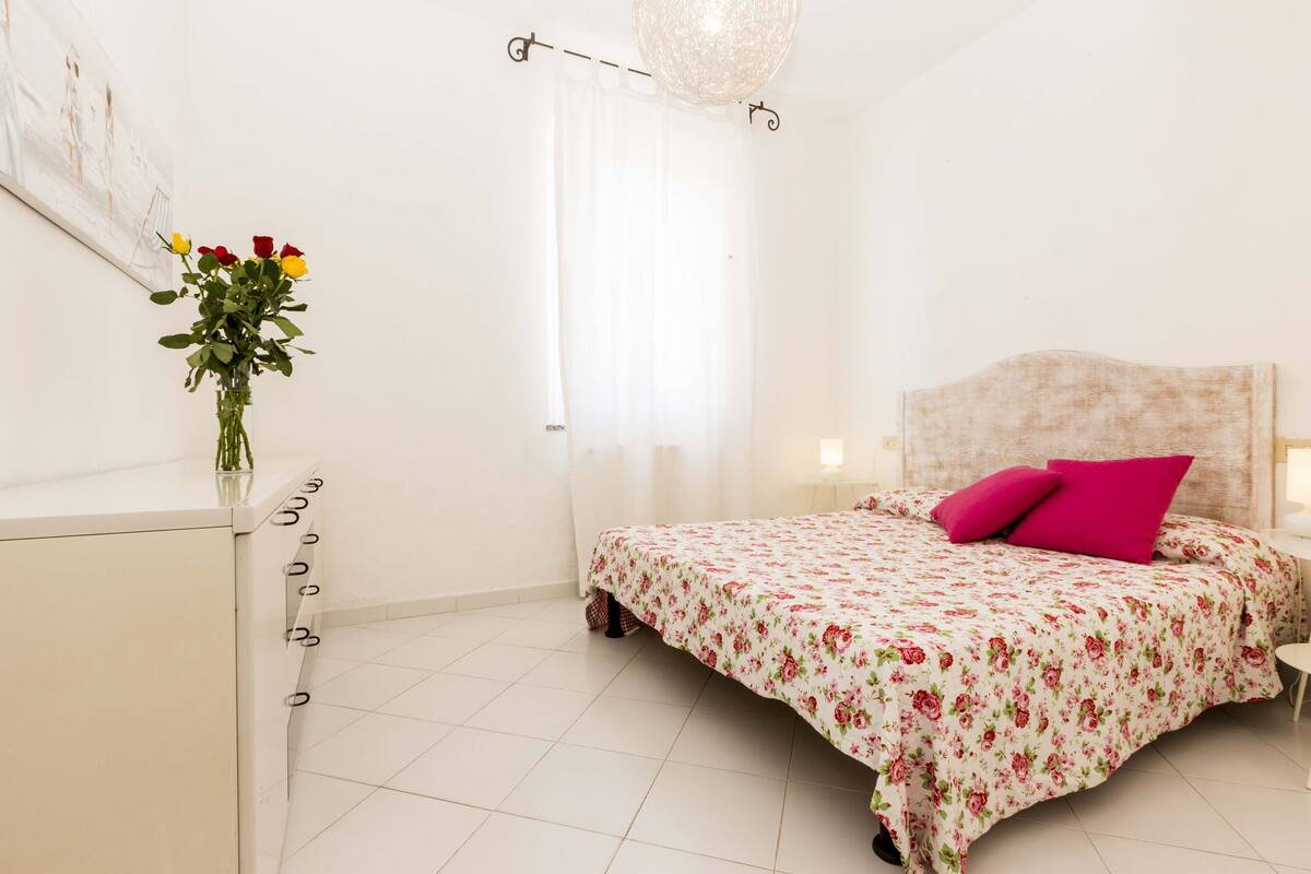 A bedroom with a bed, a nightstand, a lamp, a window with curtains, and a vase with flowers on a dresser.