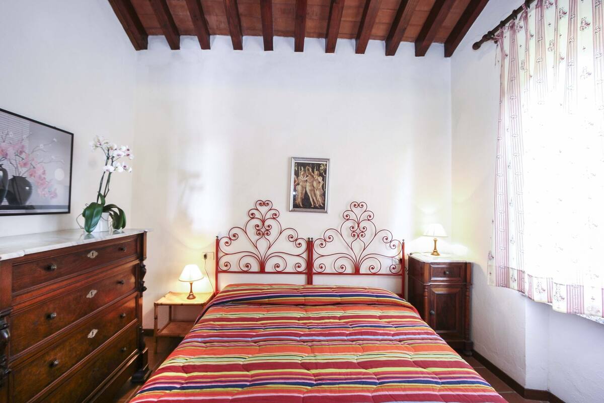 A bedroom with a wooden headboard, a colorful striped bedspread, a wooden dresser, and a small bedside table with a lamp.