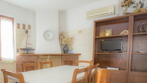Une cuisine avec des meubles de rangement en bois, une cuisinière, une table à manger avec des chaises, une horloge et un téléviseur posé sur une étagère.