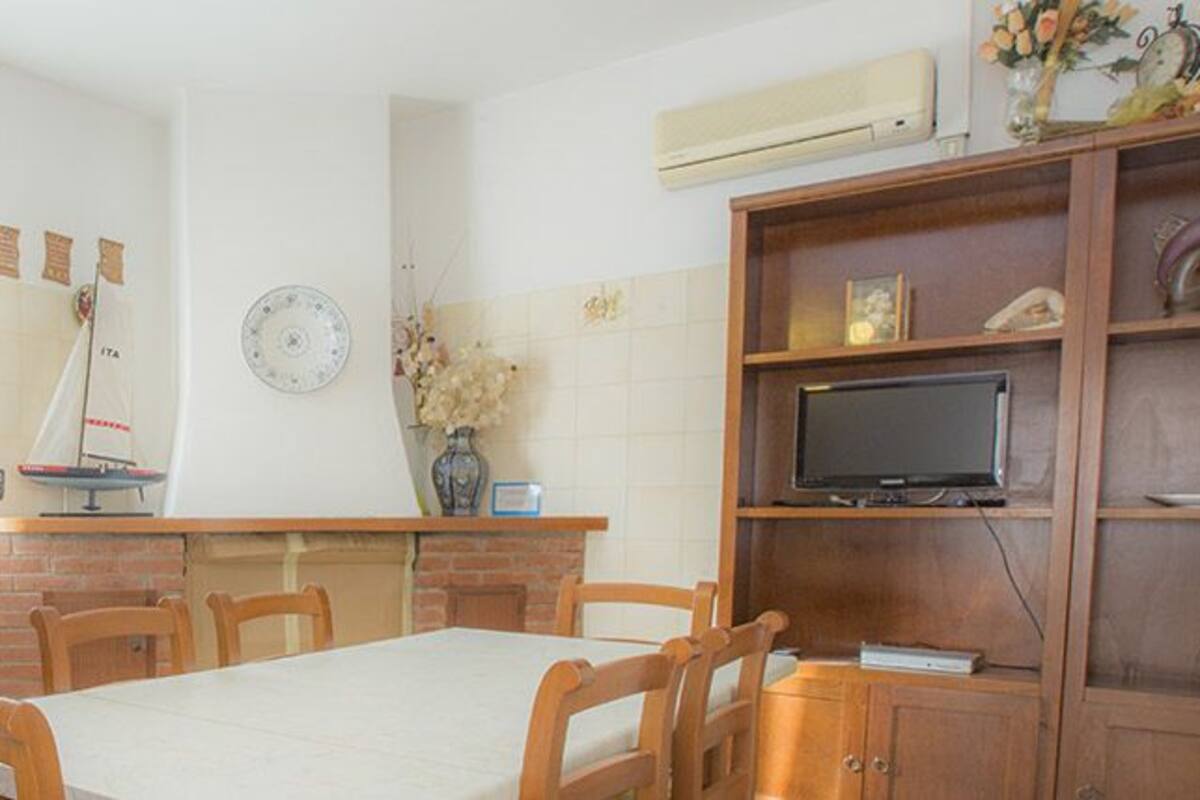 A kitchen with wooden cabinets, a stove, a dining table with chairs, a clock, and a television on a shelf.