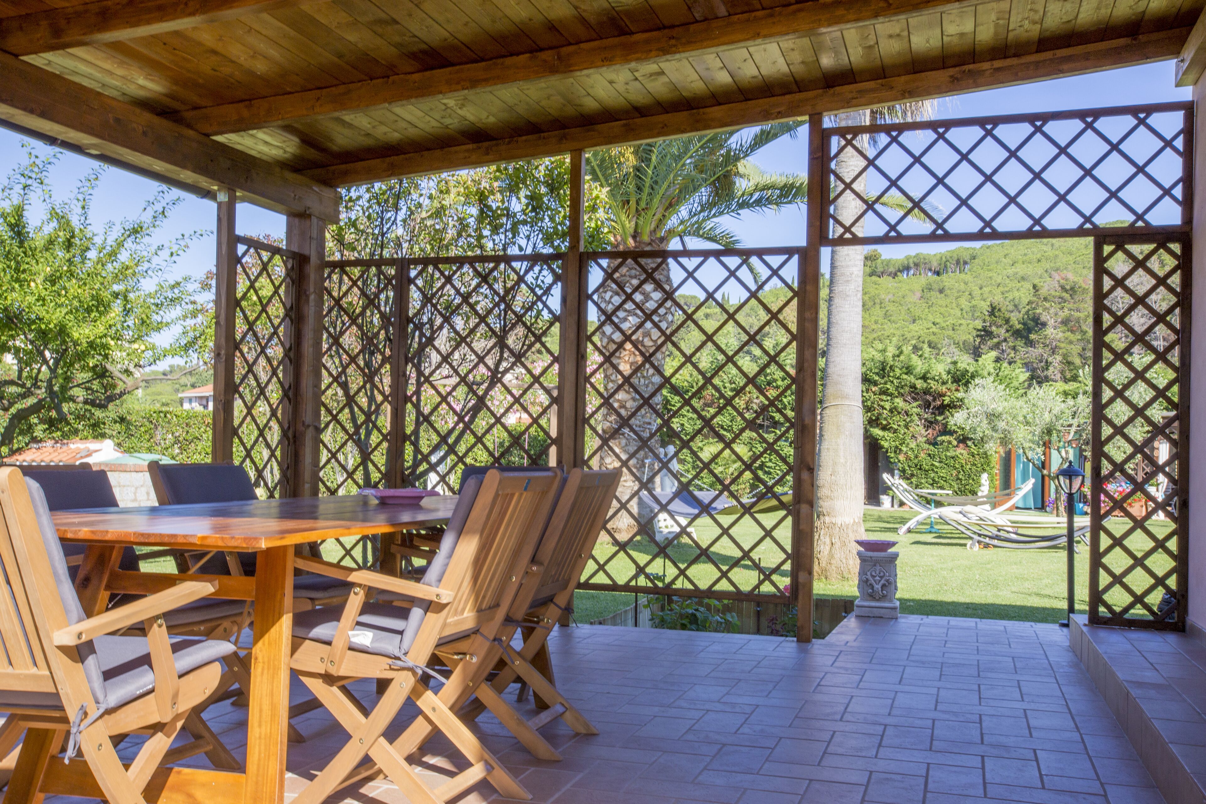 Un porche cubierto con una mesa y sillas de madera, rodeado por un enrejado y con vistas a un jardín exuberante.