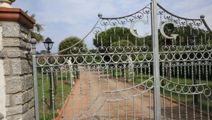 Um portão de ferro forjado com um emblema circular, dando acesso a um caminho pavimentado rodeado por vegetação.
