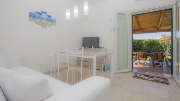 Uma sala de estar moderna com um sofá branco, uma mesa de jantar com cadeiras, uma televisão de tela plana e uma porta de vidro deslizante que dá para um pátio externo.