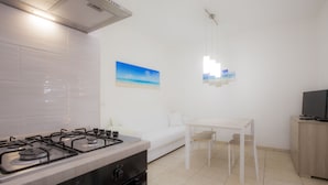 Cozinha moderna com fogão a gás, forno e painel de azulejos brancos. Há um sofá branco e uma pequena mesa de jantar com cadeiras. Um quadro de uma praia está na parede, e um lustre pendurado ilumina a mesa.
