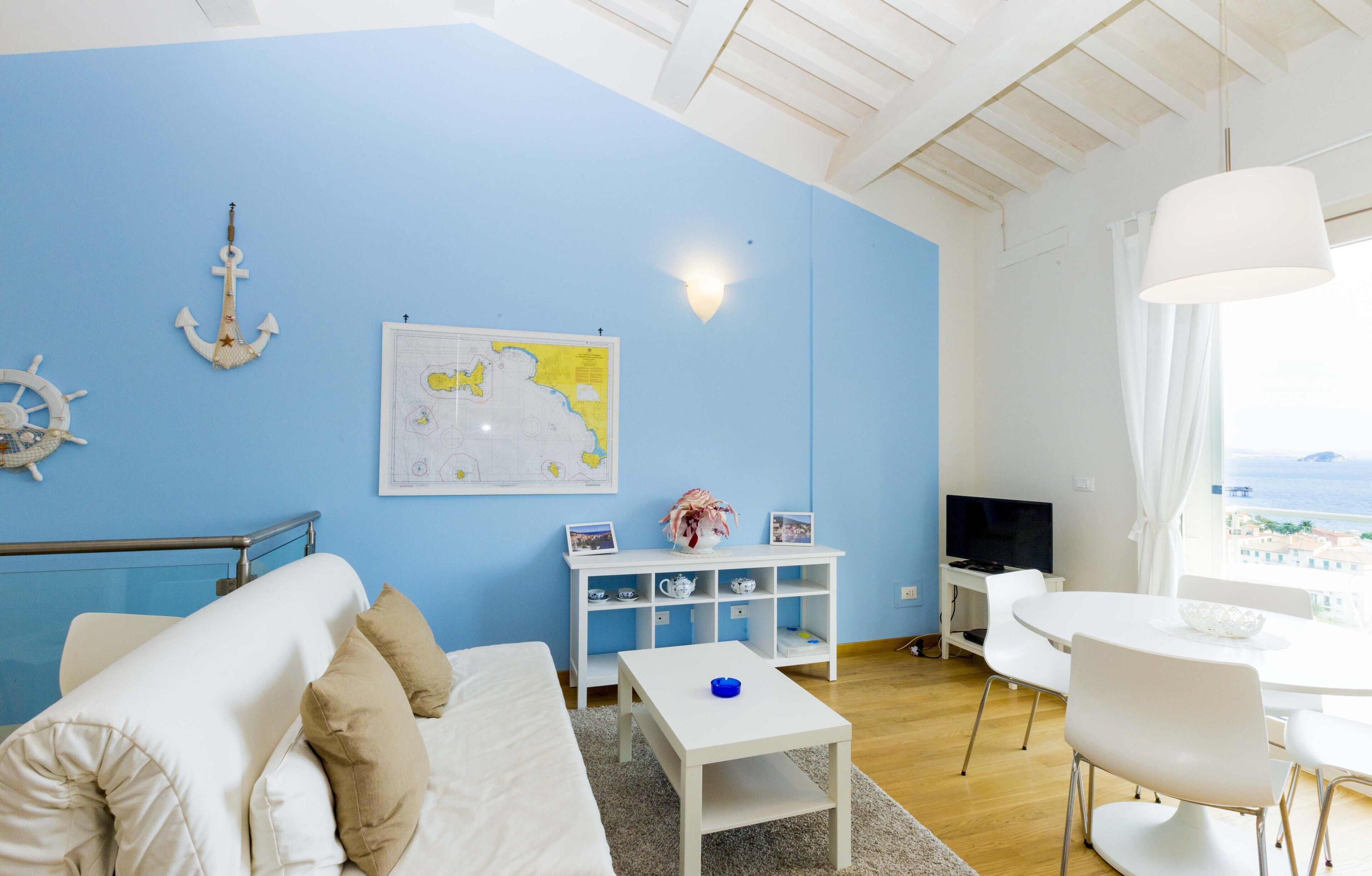 A room with a blue wall, a white sofa, a small table, a white TV stand, and a dining area with a white table and chairs.
