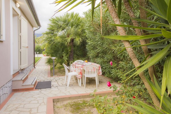 A patio with a table and chairs, surrounded by lush greenery and a building with a white exterior.