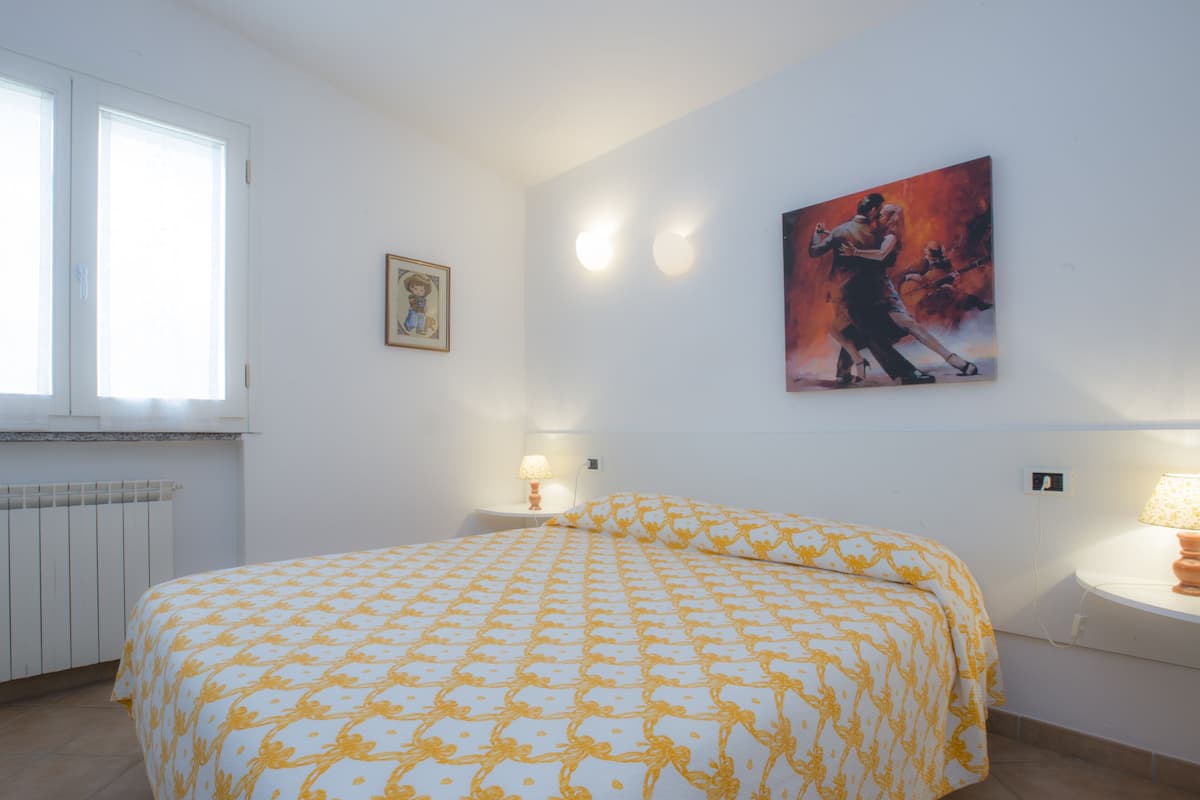 A bedroom with a bed, a nightstand, a wall-mounted lamp, a framed picture, and a window.