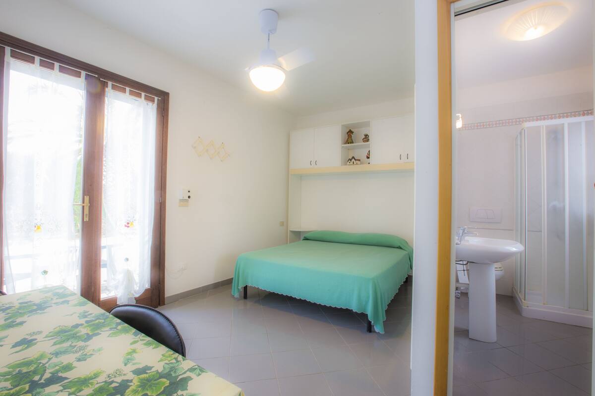 A bedroom with a bed, a chair, a ceiling fan, a window with curtains, a bathroom with a sink and toilet.