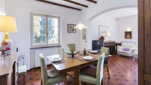 A dining area with a wooden table set for six, surrounded by chairs. There is a window with a view of greenery outside, a radiator below, and a television on a cabinet to the right. - Villa Lilla (Portoferraio)