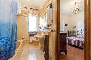 Un baño con bañera, inodoro y lavamanos. En la habitación contigua hay una cama con colcha a cuadros.
