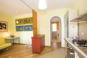 A compact kitchen with a gas stove, a refrigerator, and a dining area with a yellow sofa and a small table. - Baia Fiorita (Porto Azzurro)