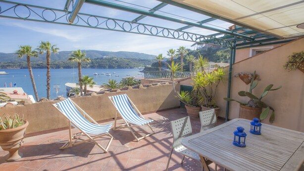 A terrace with a table and two striped chairs, overlooking a body of water with boats and palm trees. - Terrazza Ferdinando (Porto Azzurro)
