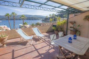 Une terrasse avec une table et deux chaises à rayures, donnant sur une étendue d’eau où l’on peut voir des bateaux et des palmiers.