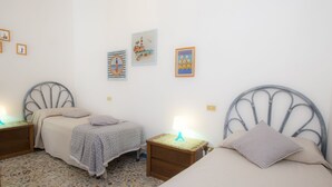 Un dormitorio con dos camas, una mesita de noche pequeña con lámpara y suelo de baldosas.