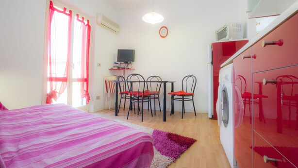 Um quarto com uma cama, uma escrivaninha com computador, uma máquina de lavar e um espaço de jantar com cadeiras.