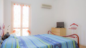 Quarto com cama, escrivaninha, cadeira, televisão e janela com cortinas.