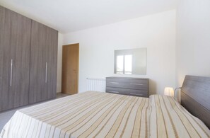 Quarto com um guarda-roupa espaçoso, uma mesa de cabeceira com abajur e uma cama com roupa de cama listrada.