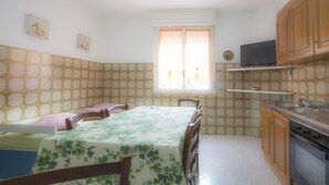 A dining area with a table set for four, a kitchen with wooden cabinets and a stove, and a window with a curtain. - Appartameno Mediterraneo (Campo nell'Elba)