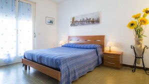 A bedroom with a bed, bedside tables, a nightstand, a window with curtains, a wall-mounted painting, and a vase with sunflowers. - Appartameno Mediterraneo (Campo nell'Elba)