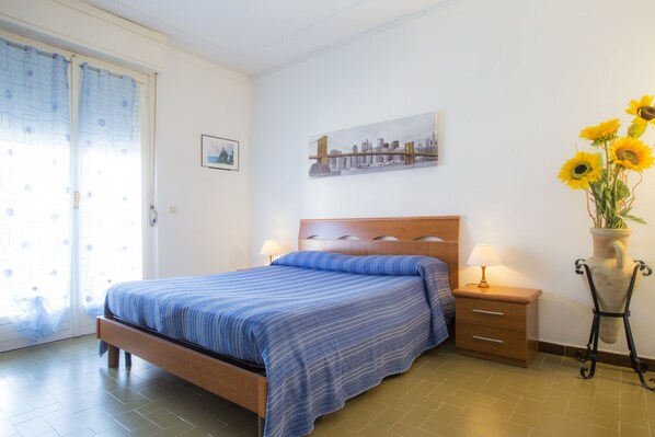 A bedroom with a bed, bedside tables, a nightstand, a window with curtains, a wall-mounted painting, and a vase with sunflowers.