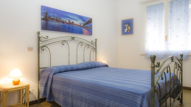 A bedroom with a metal bed frame, a blue-striped bedspread, a nightstand with a lamp, and a cityscape painting on the wall. - Appartameno Mediterraneo (Campo nell'Elba)