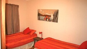 A room with two beds, each with red bedding and pillows, a small bedside table with a lamp, and a painting on the wall depicting a landscape with birds and trees. - Nestled Atop the Lane - Pukawa Bay Holiday Home (Pukawa Bay)