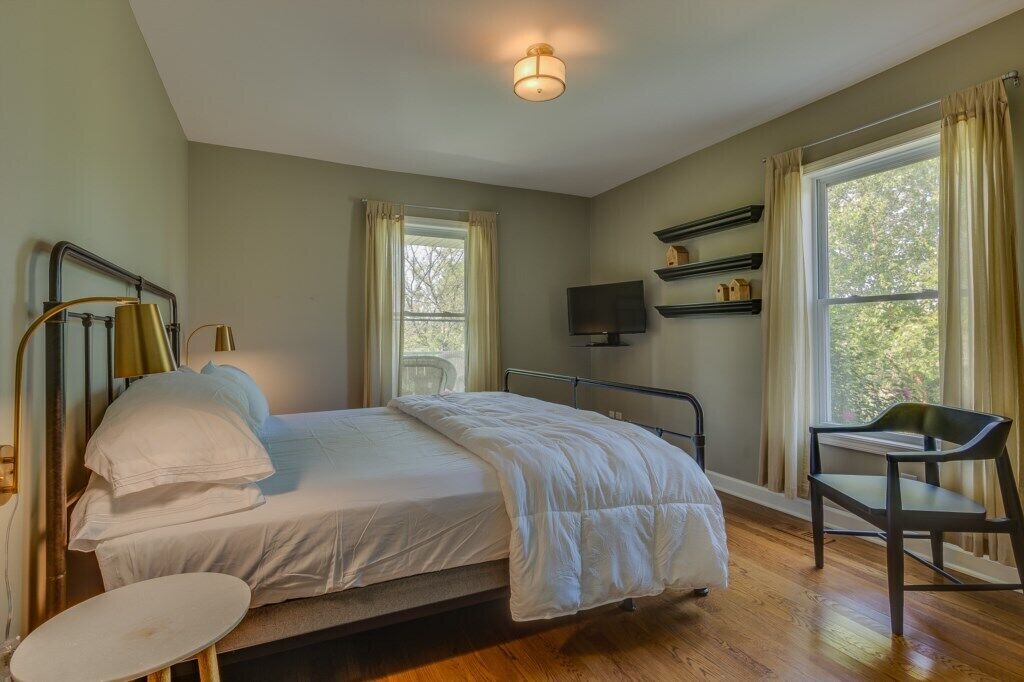 A bedroom with a bed, a chair, a TV, and a window with curtains.