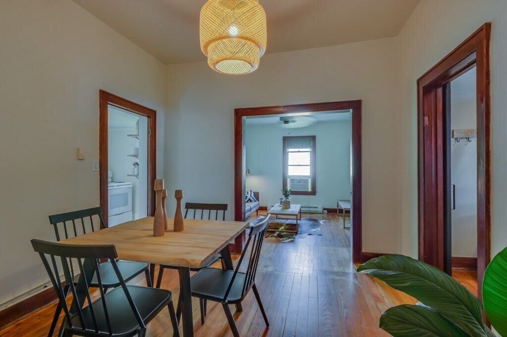 A dining area with a wooden table and chairs, a hanging light fixture, and a view into another room with a window.